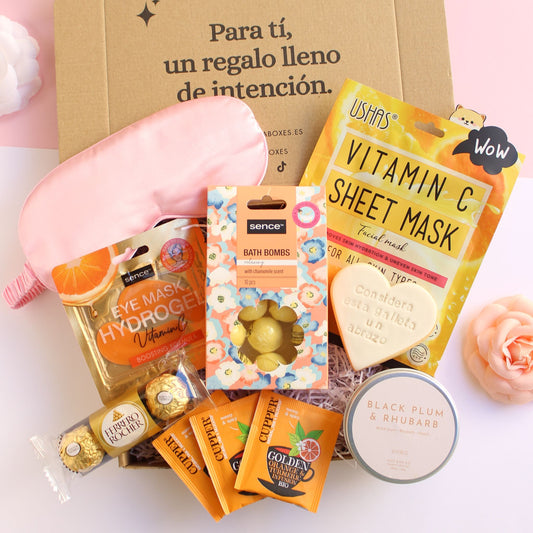 Caja regalo treat box para nueva mamá o madre primeriza que contiene antifaz satinado de color rosa, parches de hidrogel para ojos, tres bombones ferrero rocher, diez mini bombas de baño con esencia de camomila, bolsitas de infusión orgánica, vela en lata de viaje con aroma de ciruela negra y ruibarbo, galleta de mantequilla con forma de corazón y el texto considera esta galleta un abrazo impreso y mascarilla facial enriquecida con vitamina c.