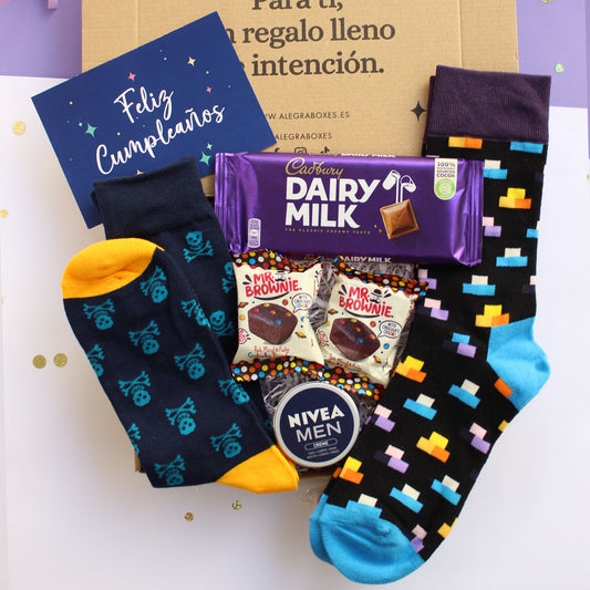 Caja regalo treat box de cumpleaños para hombre que contiene unos calcetines con diseño de piezas de tetris, otros calcetines azul marino con diseño de calaveras, dos mini brownies de mr. brownie, lata de crema nivea men, tableta de chocolate dairy milk de la marca cadbury y una tarjeta de felicitación con el texto feliz cumpleaños.