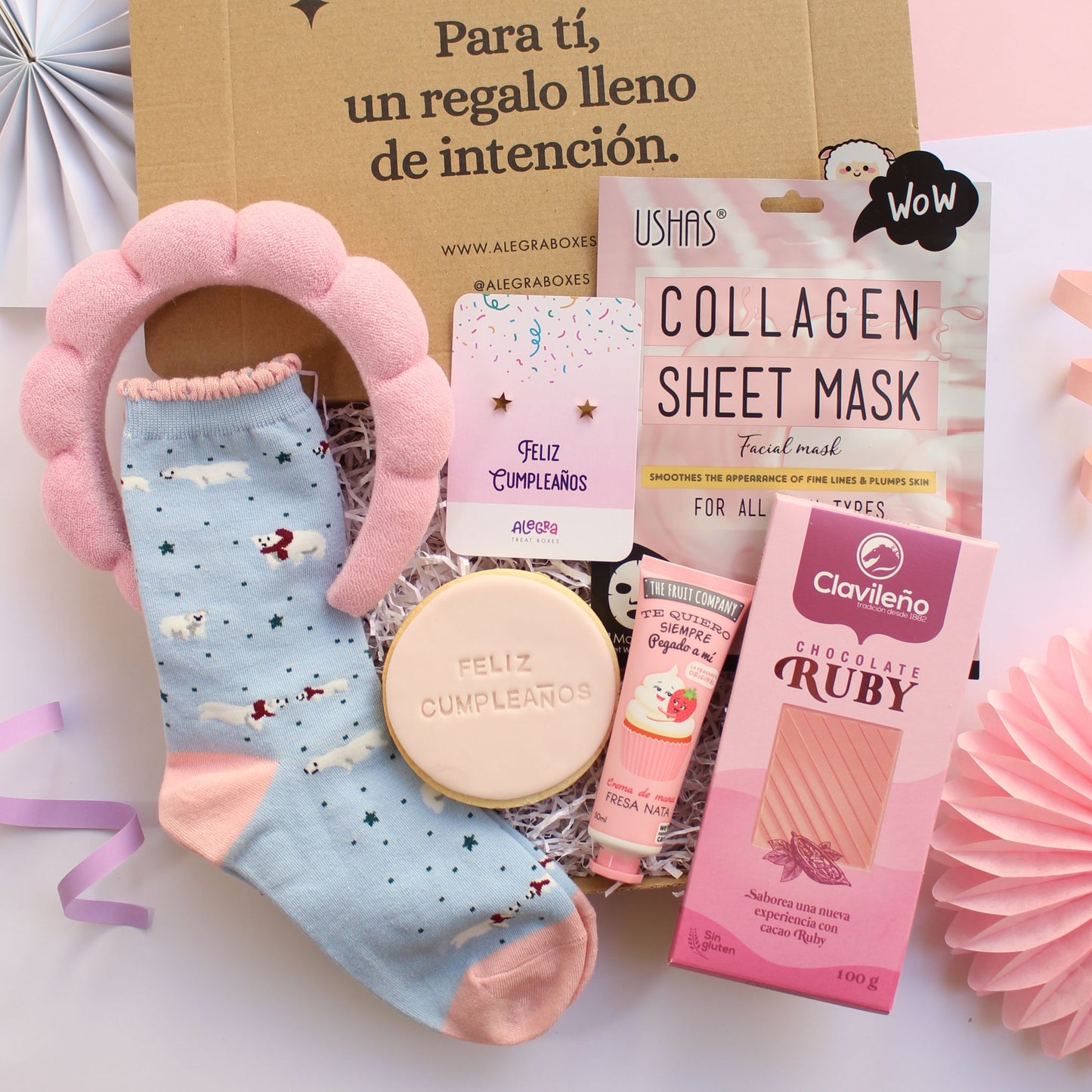 Caja regalo treat box de cumpleaños para mujer que incluye unos calcetines celeste y rosa con diseño de osos polares, diadema de esponja rosa para maquillaje, galleta de mantequilla redonda con el texto feliz cumpleaños impreso, pendientes de acero dorado con forma de estrella en cartulina con el texto feliz cumpleaños, mascarilla facial de colágeno, tableta de chocolate ruby y crema de manos fresa nata.