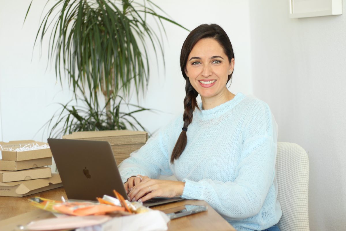 Claudia, creadora de Alegra Treat Boxes posando sonriente mientras trabaja en su portátil, rodeada de cajas de envío y algunos productos.