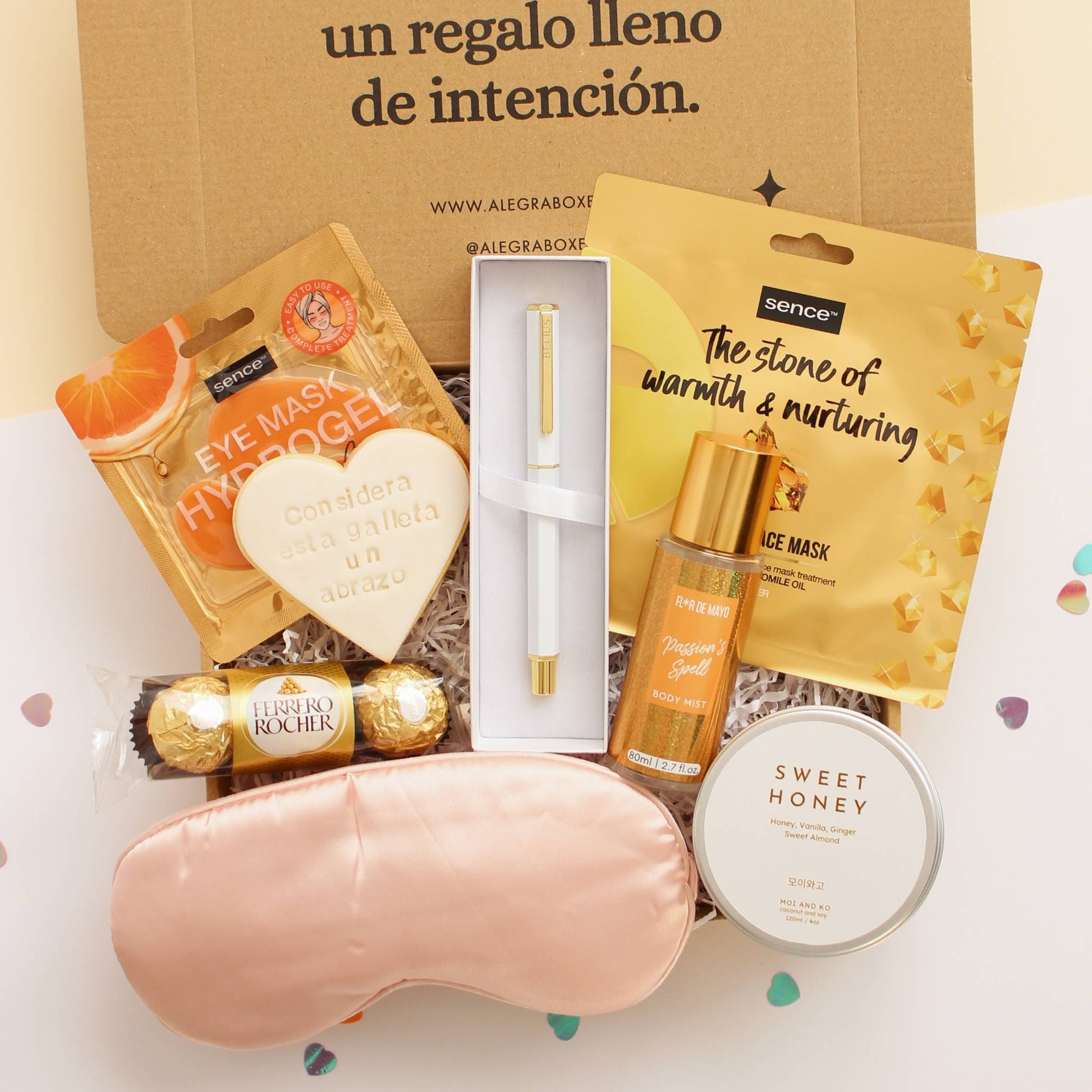 Caja regalo treat box para novias que incluye parches para ojos de hidrogel, galleta de mantequilla en forma de corazón y mensaje considera esta galleta un abrazo impreso, tres bombones de Ferrero Rocher, antifaz satinado de color rosa, elegante bolígrafo de cuerpo hexagonal en color blanco con detalles dorados, frasco de bruma corporal, vela en lata de viaje con fragancia sweet honey y mascarilla facial nutritiva.