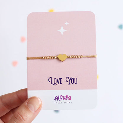 Pulsera de hilo color dorado con corazón de acero dorado en cartulina con la leyenda Love You