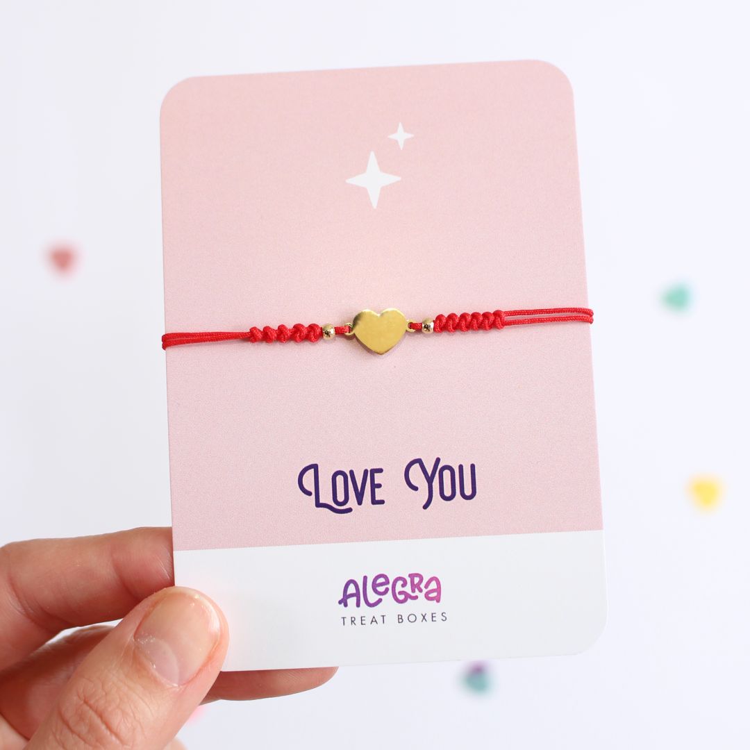 Pulsera de hilo color rojo con corazón de acero dorado en cartulina con la leyenda Love You