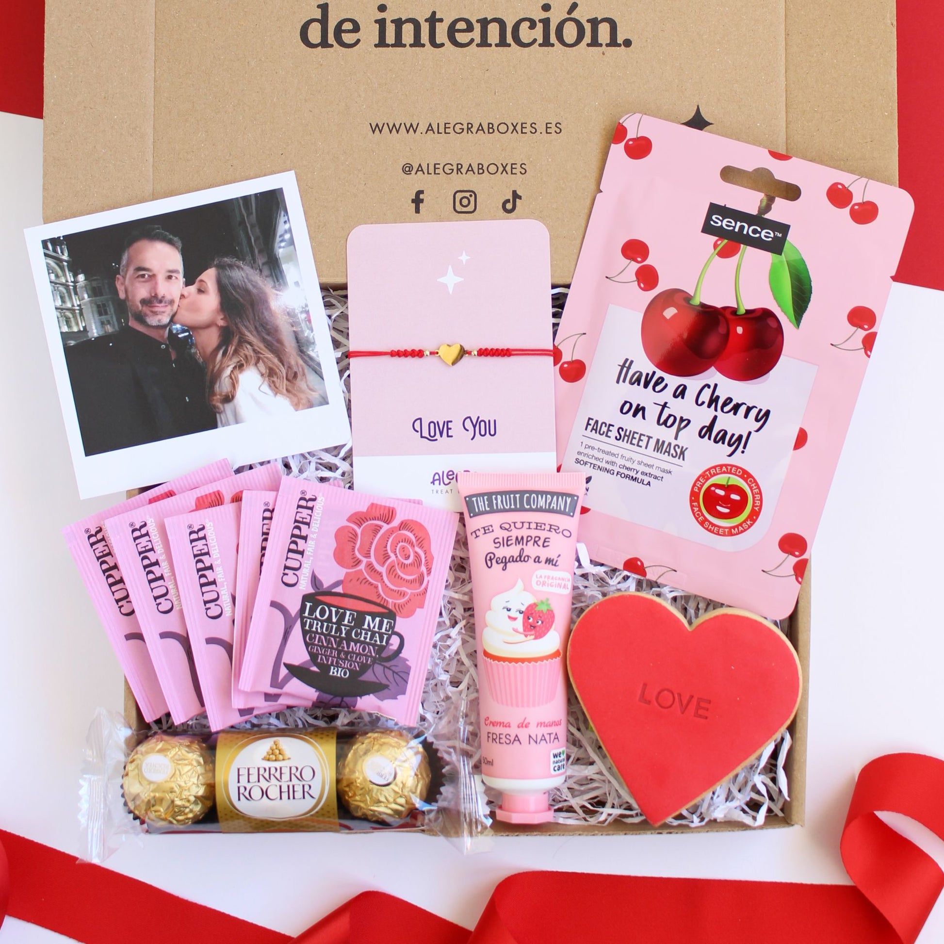 Caja regalo treat box para mujer the love ox que incluye fotografía personalizada Polaroid, cinco sobres de infusión orgánica, tres bombones Ferrero Rocher, crema de manos fresa nata, galleta de mantequilla roja con forma de corazón y la palabra love impresa, mascarilla facial con efecto suavizante y pulsera de hilo rojo con corazón de acero dorado en cartulina con el texto Love you.